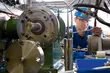 Techniker mit Schutzhelm und Schutzbrille arbeitet an einer komplexen industriellen Anlage mit Rohren, Dichtungssystemen und Messinstrumenten.